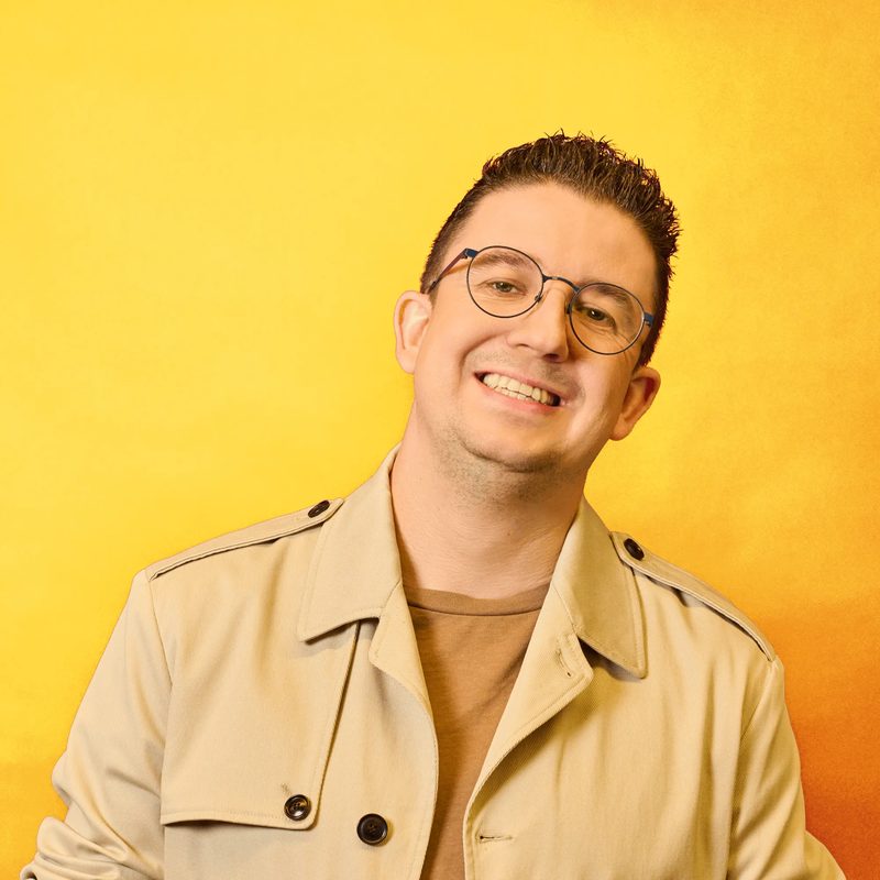 Mark van Gils, smiling, wearing round glasses and a beige jacket in front of a yellow background